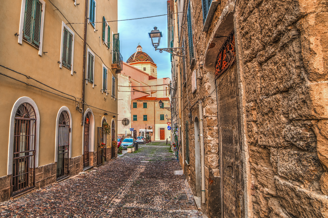 Kopfsteinpflastergasse in der Altstadt von Alghero mit hohen Häusern, bunten Fassaden und Blick auf eine Kirche mit Kuppel.