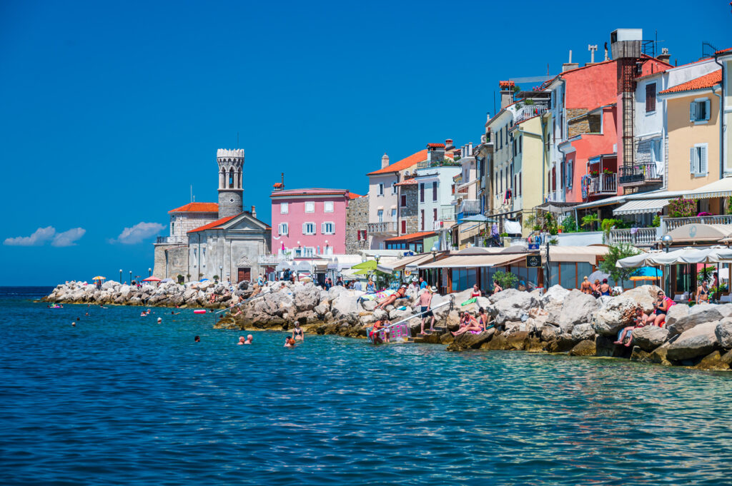 Bunt gestrichene Häuser und die Kirche am Meer in Piran in Slowenien mit vielen Badenden auf den Felsen.