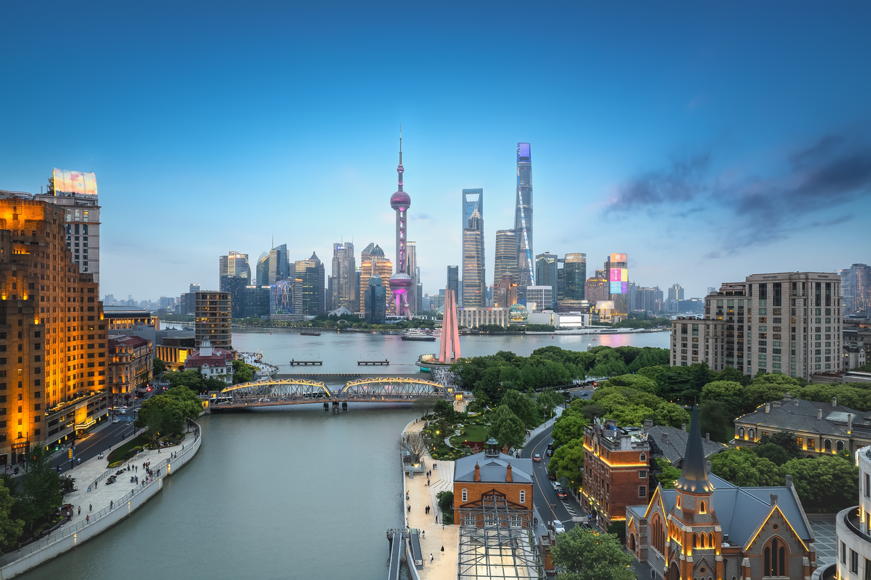 Blick über den Huangpu-Fluss auf die erleuchtete Skyline von Shanghai mit dem Oriental Pearl Tower und anderen Hochhäusern bei einsetzender Dämmerung.