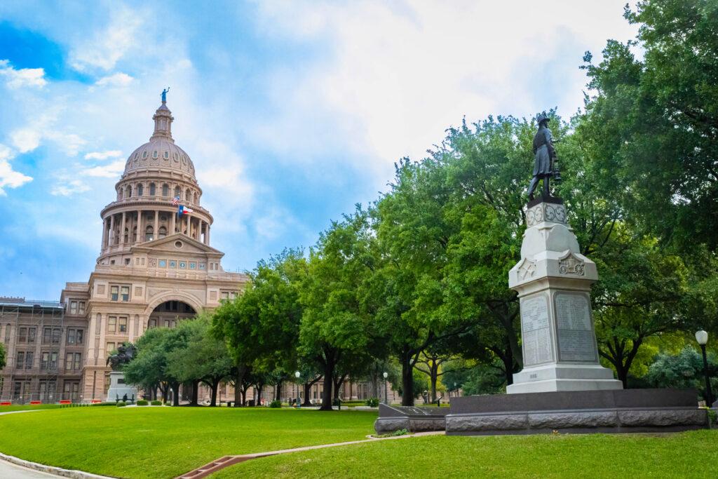 Die Kuppel des Texas State Capitol erhebt sich hinter grünen Bäumen und einem Denkmal im Park in Austin, Texas.