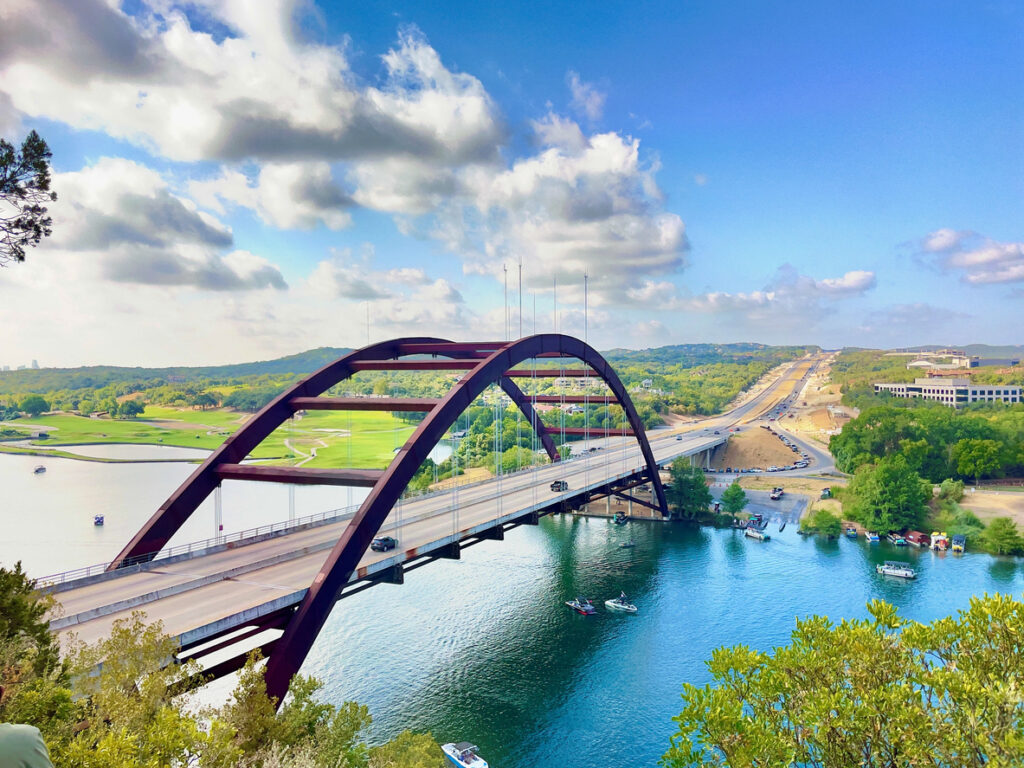 Luftaufnahme der geschwungenen Pennybacker Bridge über einem blaugrünen Fluss mit sanften Hügeln und Straßen rund um Austin, Texas.