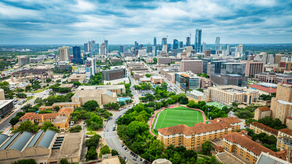 Luftaufnahme von Austin, Texas, mit Universitätsgebäuden und einem Sportfeld im Vordergrund sowie der hoch aufragenden Downtown im Hintergrund.