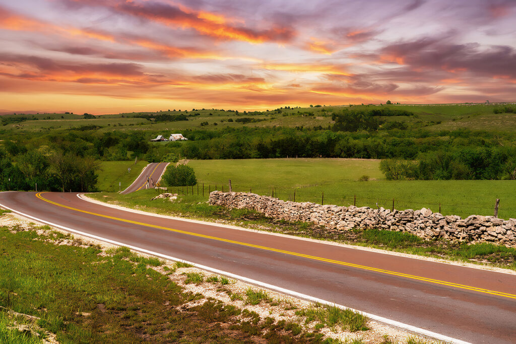 Geschwungene Landstraße mit Steinmauer in den grünen Flint Hills bei farbigem Sonnenuntergangshimmel.