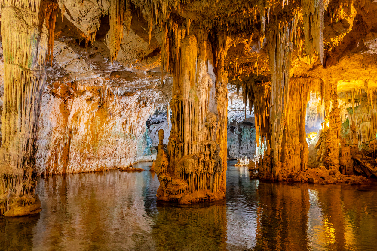 Im Inneren einer Tropfsteinhöhle bei Alghero hängen stalaktiten von der Decke und spiegeln sich im Wasser.