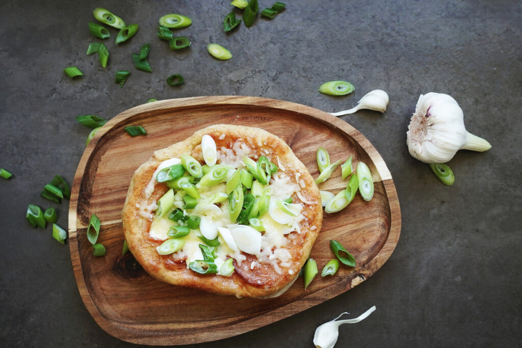 Lángos mit Käse und Frühlingszwiebeln auf einem Holzbrett, daneben frische Knoblauchknollen.