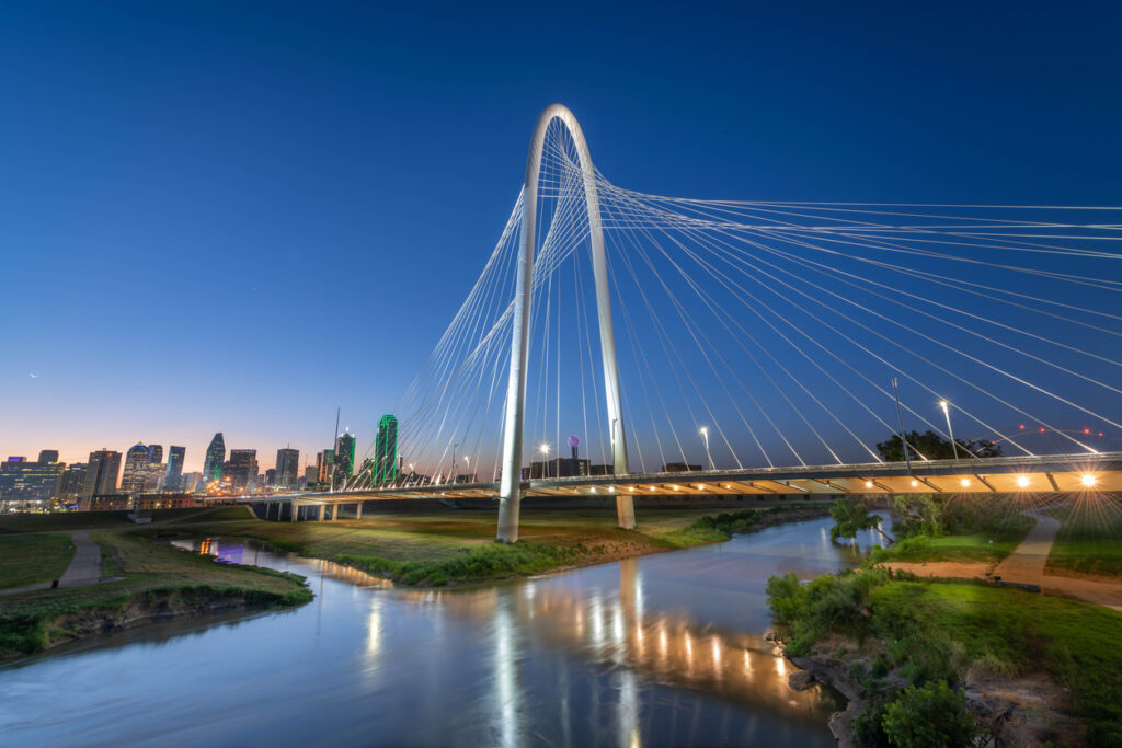 Die Margaret Hunt Hill Bridge spannt sich bei blauem Abendhimmel über den Trinity River, im Hintergrund leuchtet die Skyline von Dallas, Texas.