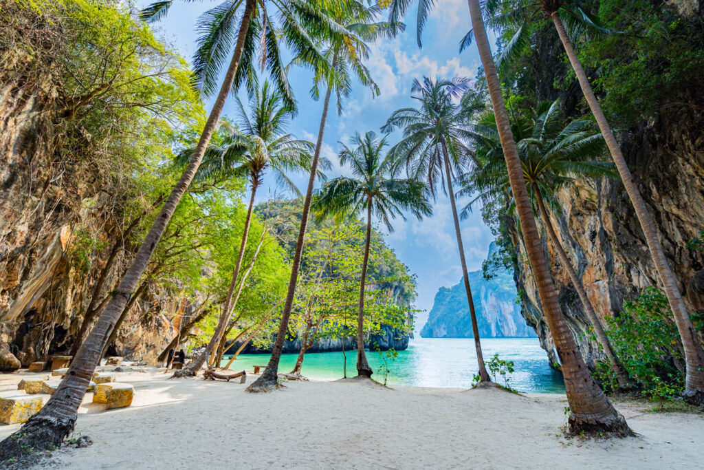 Palmenbestandener Sandstrand in einer von Felsen umrahmten türkisblauen Bucht bei Krabi in Thailand.