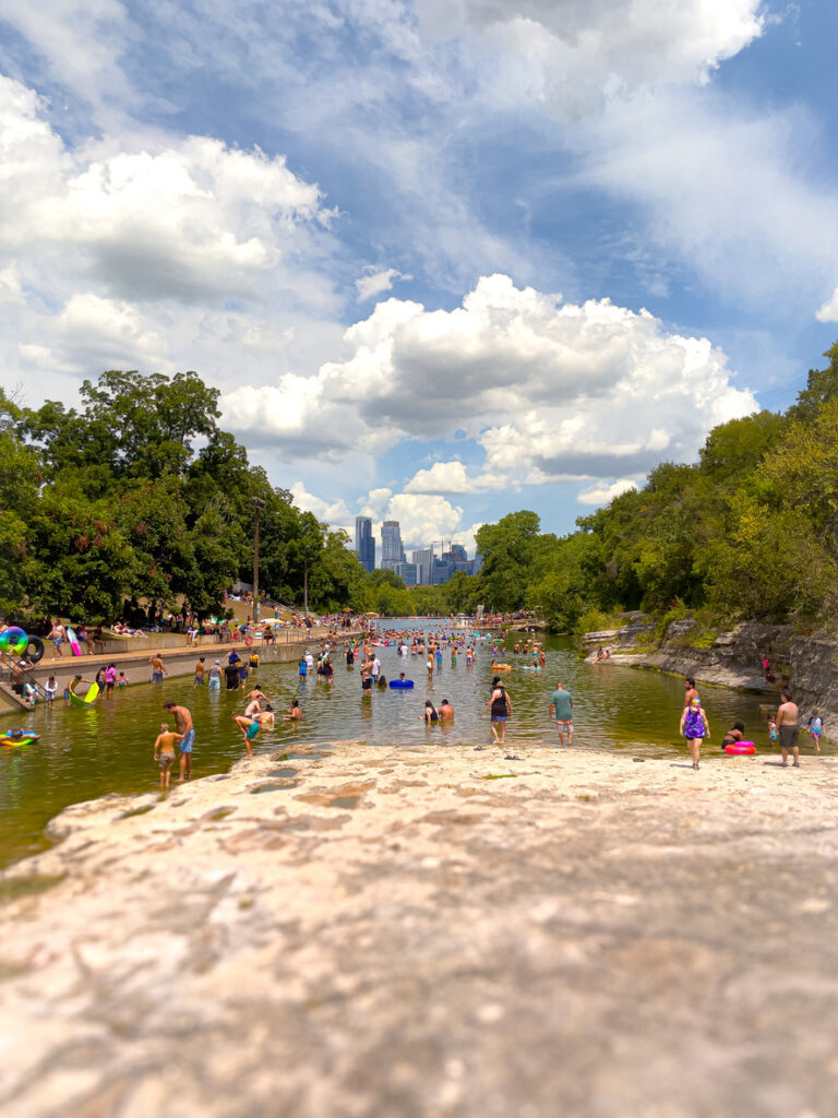 Viele Menschen baden und spielen in einem natürlichen Schwimmbecken, im Hintergrund ist zwischen den Bäumen die Skyline von Austin, Texas zu sehen.