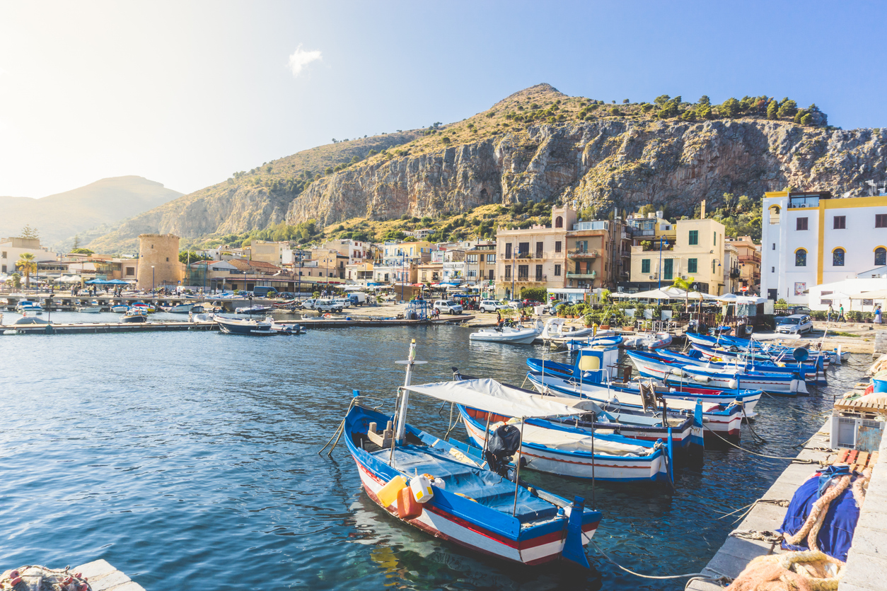 Fischerboote im Hafen bei Palermo vor Felsen und Häusern am Ufer.