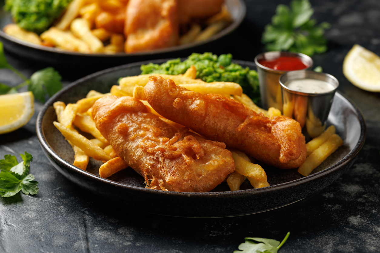 Ein Teller mit goldbraunen Fish and Chips, Erbsenpüree und Dipsauce steht auf einem dunklen Tisch.