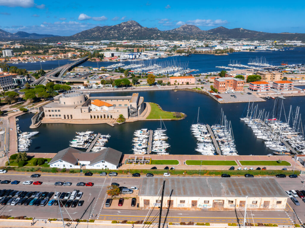 Luftaufnahme von Olbia mit Yachthafen, Brücken, modernen Gebäuden und Bergen im Hintergrund.