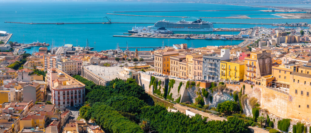 Luftaufnahme von Cagliari mit Altstadt, Hafenbecken und Kreuzfahrtschiff in der tiefblauen Bucht.