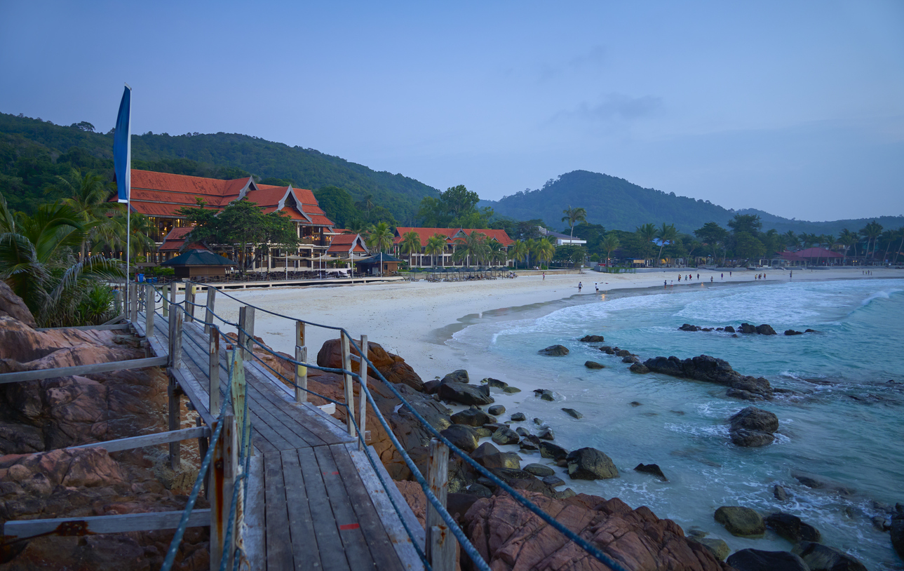 Ein Holzsteg führt auf Redang entlang der Felsen zu einem langen Sandstrand mit Resorts im Hintergrund.