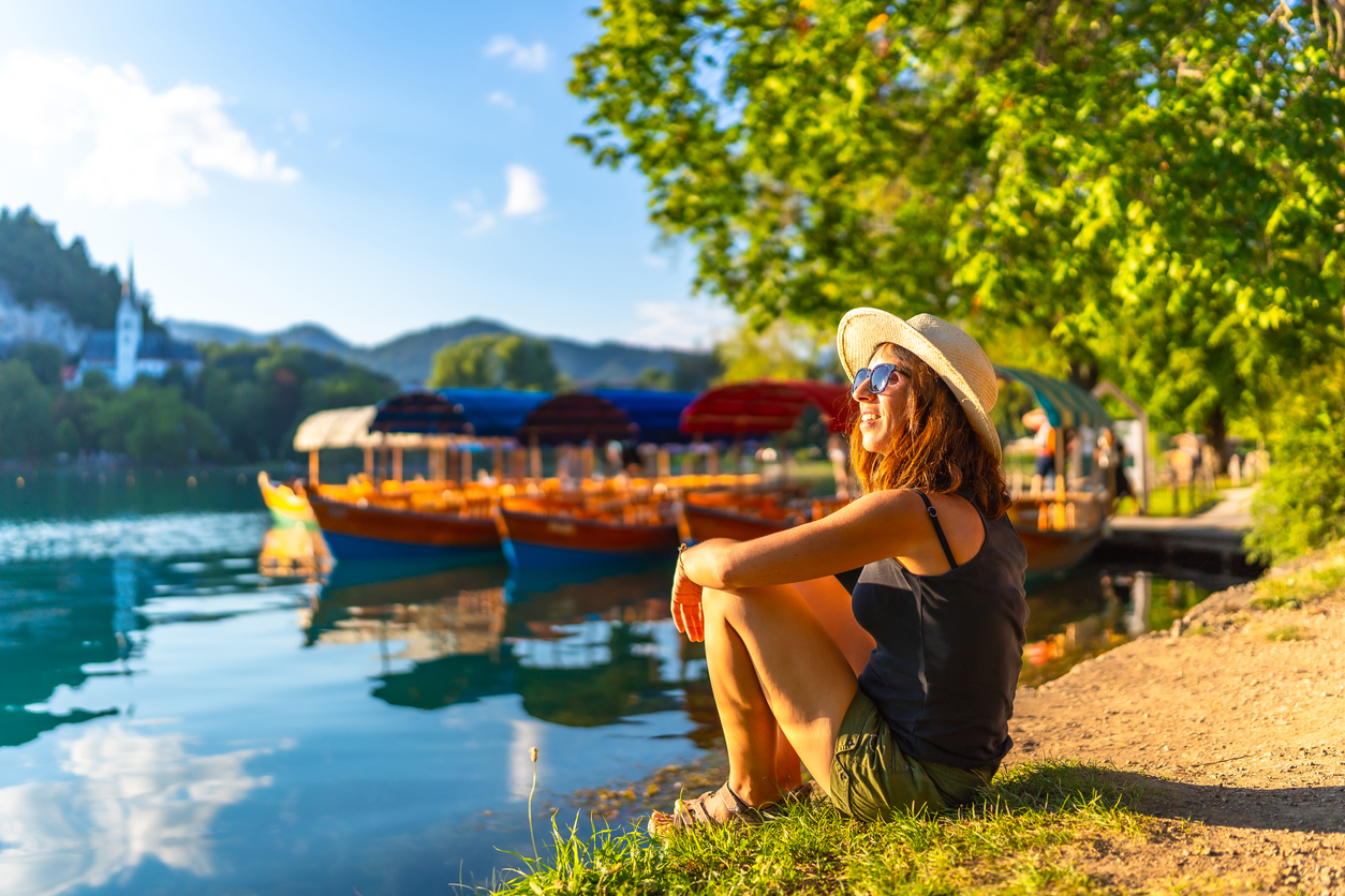 Junge Frau sitzt entspannt am Ufer vom Bleder See in Bled und blickt auf die traditionellen Holzboote.