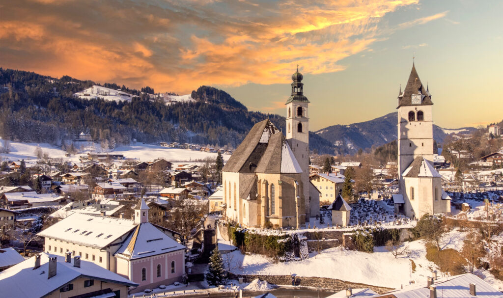 Blick auf die verschneite Altstadt von Kitzbühel mit Kirche und Bergen im Abendlicht.