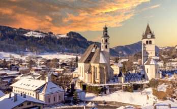 Blick auf die verschneite Altstadt von Kitzbühel mit Kirche und Bergen im Abendlicht.