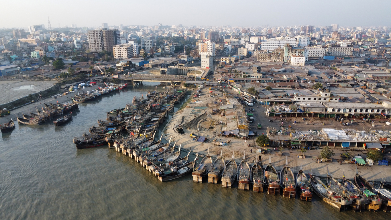 Luftaufnahme des Hafens von Chittagong / Chattogram mit dicht aneinandergereihten Fischerbooten und Stadtpanorama im Hintergrund.