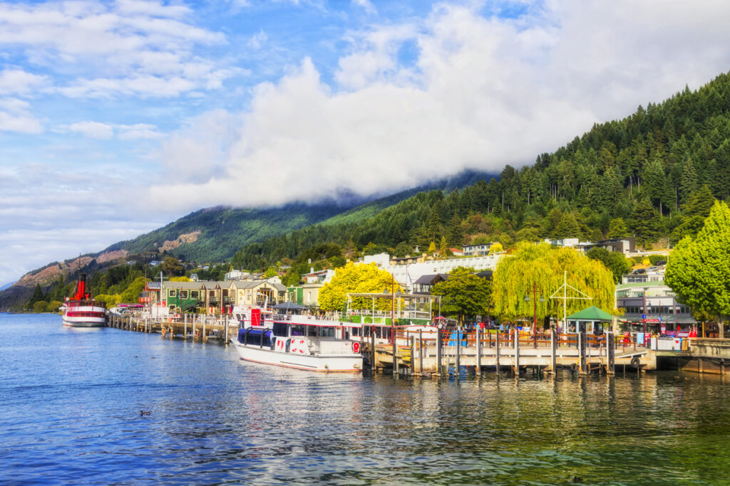 Boote und Stege an der Uferpromenade von Queenstown vor bewaldeten Hügeln und tief hängenden Wolken.