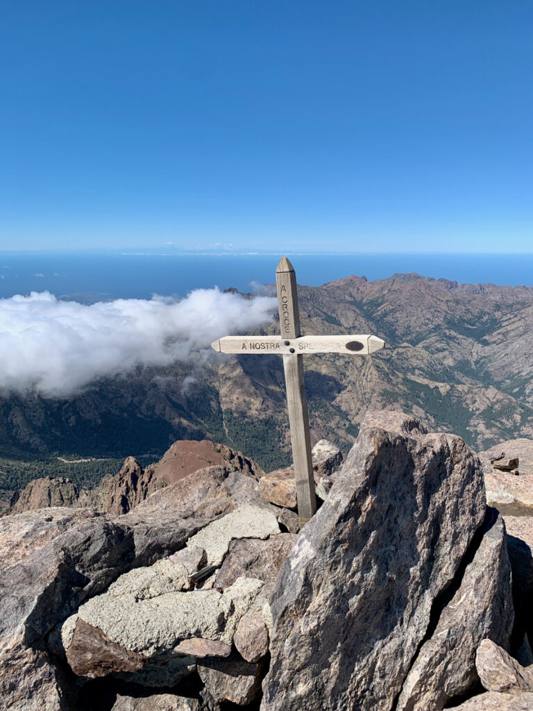 Ein hölzernes Kreuz steht auf felsigem Gipfelgelände am Monte Cinto, dahinter liegen Berge, Wolken und der blaue Horizont.