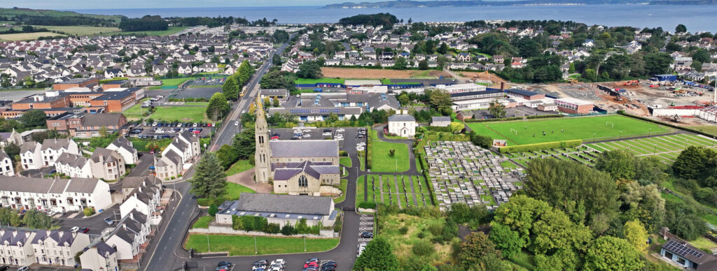 Luftaufnahme von Ballycastle mit Kirche, Wohnvierteln, grünen Feldern und dem Meer im Hintergrund.