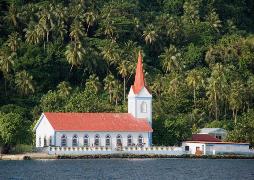 Weiße Kirche mit rotem Dach steht direkt am Meer in Französisch-Polynesien vor dichter Palmenkulisse.