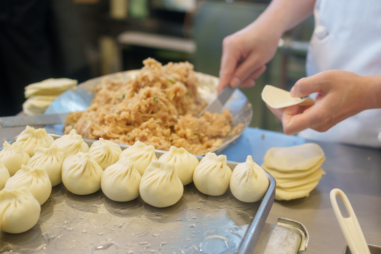 Hände formen Dumpling-Teig in einer Garküche in Shanghai, während gefüllte Teigtaschen in Reihen auf einem Metallblech liegen.