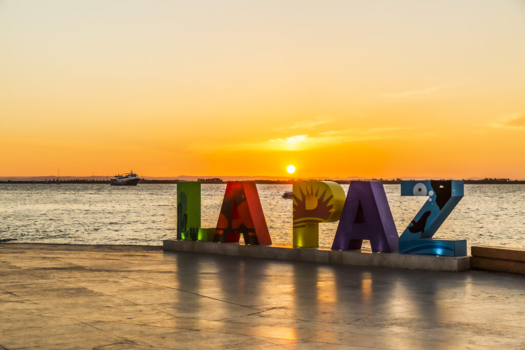 Bunte LA PAZ Buchstaben stehen an einer Uferpromenade vor der untergehenden Sonne.