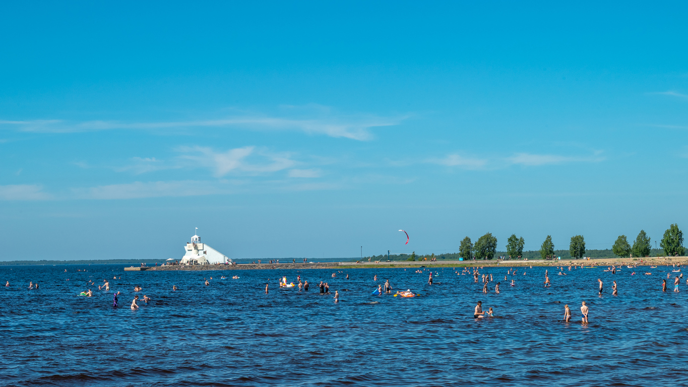 Viele Menschen baden im flachen Wasser vor einer kleinen weißen Leuchtfeueranlage bei Oulu.