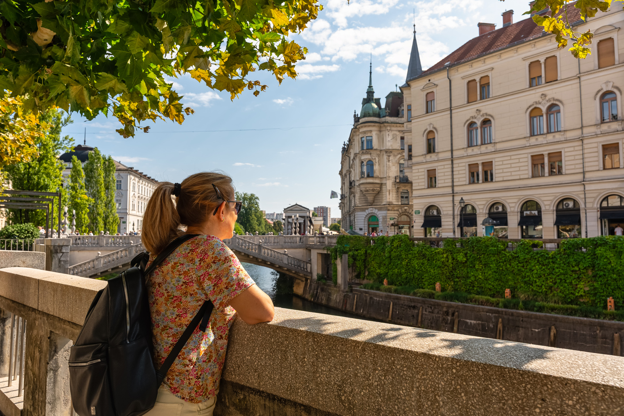 Frau mit Rucksack blickt in Ljubljana über die Ljubljanica auf Brücke und Altstadt.