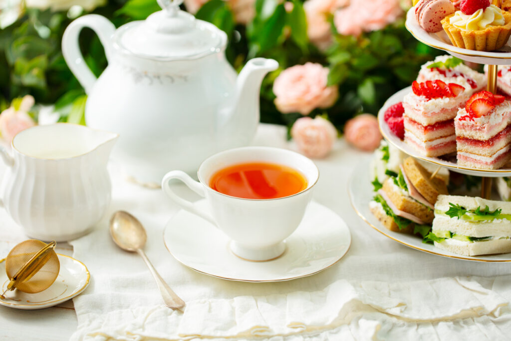 Gedeckter Tisch mit Teetasse, Kanne, Milchkrug und Etagere voller Sandwiches und Süßspeisen für einen Afternoon Tea.