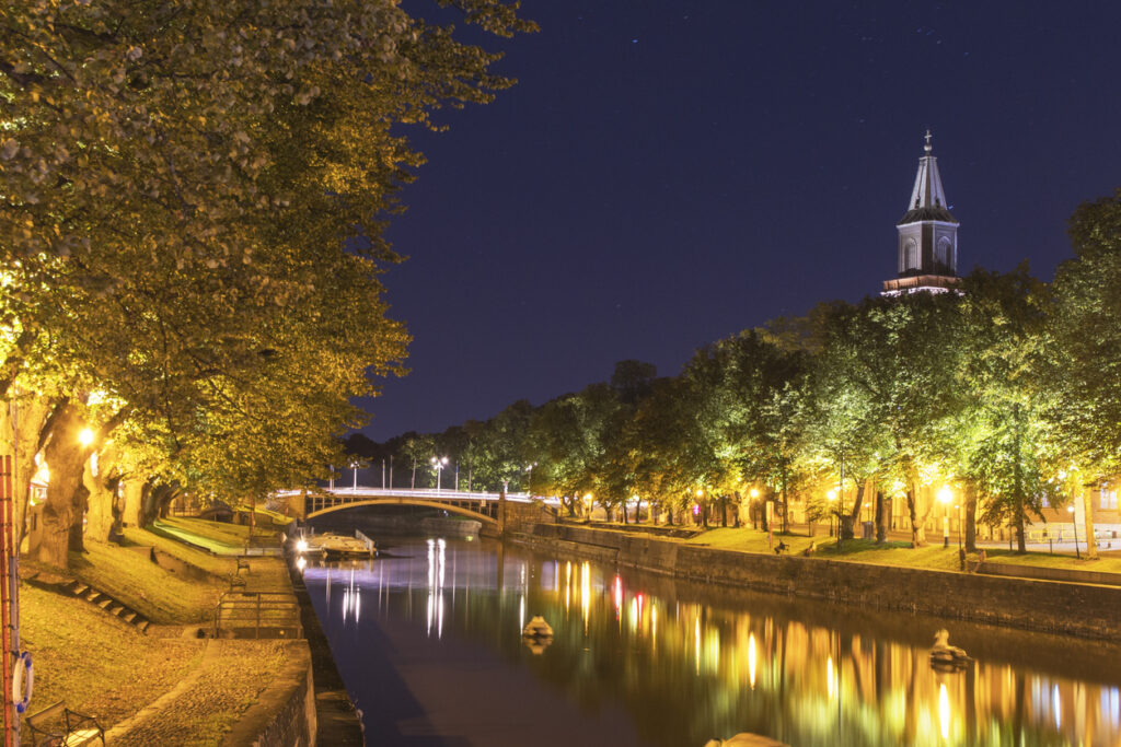 Beleuchtete Uferpromenade mit Kirchturm spiegelt sich im Fluss in Turku.