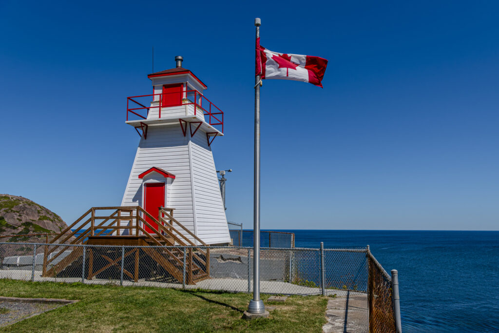 Weißer Leuchtturm mit roter Tür und wehender Kanada-Flagge an der Küste von Neufundland.