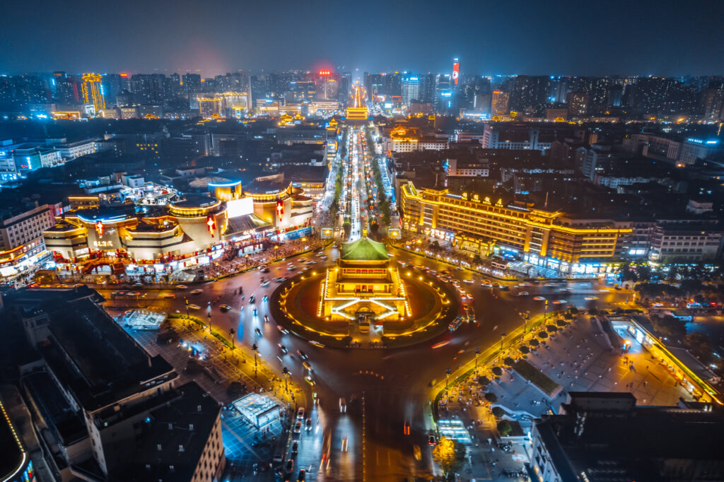 Nachtaufnahme des beleuchteten Glockenturms von Xi’an in einem Kreisverkehr, von hell erleuchteten Straßen und Hochhäusern umgeben.