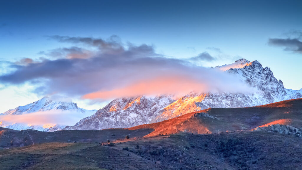 Der schneebedeckte Monte Cinto wird von rosa Wolken und warmem Sonnenlicht über dunklen Hängen eingerahmt.