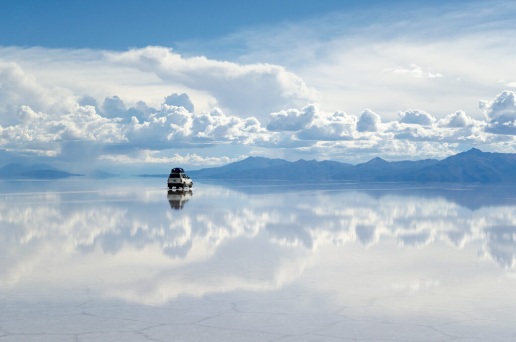 Ein Auto fährt über den spiegelglatten Salzsee Salar de Uyuni bei Uyuni unter dramatischen Wolken.