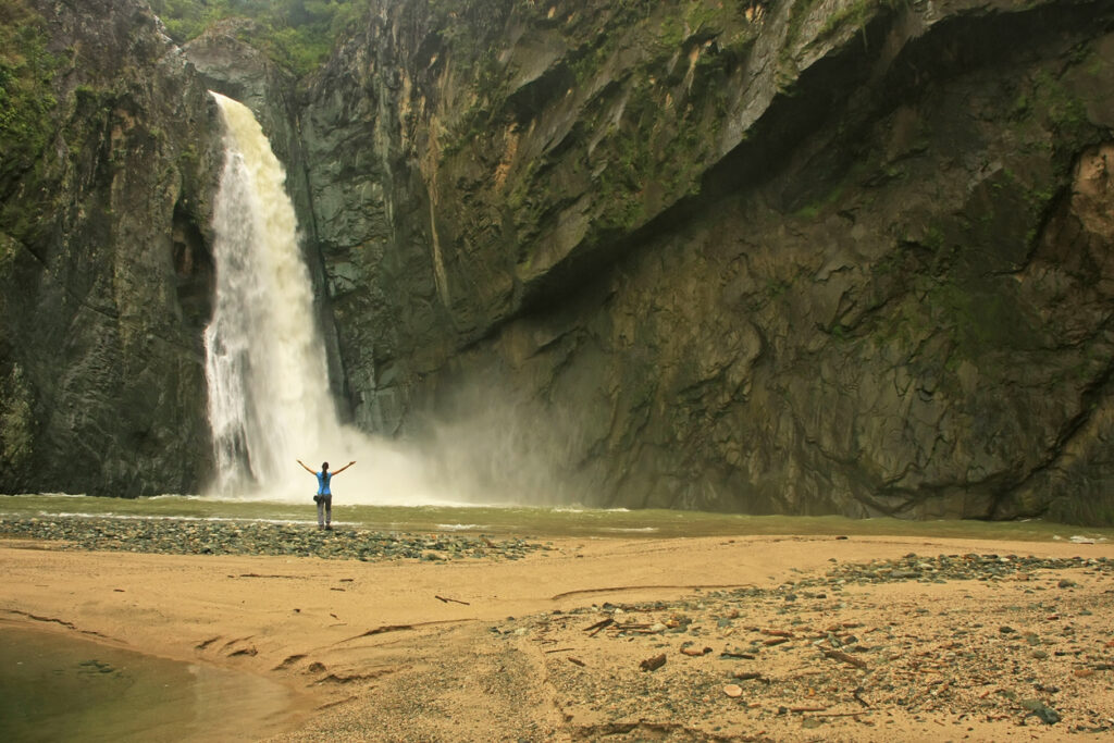 Eine Person steht vor einem hohen Wasserfall in einer felsigen Schlucht bei Jarabacoa.
