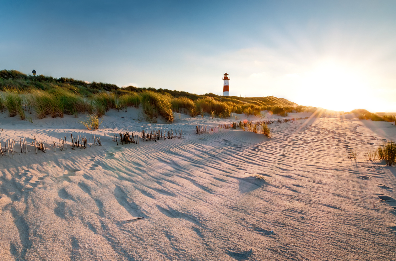 Ein Leuchtturm steht in den Dünen auf Sylt, während die Sonne tief über dem Sand scheint.