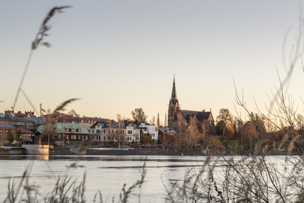 Blick über den Fluss auf Umeå mit Kirchturm und Häusern bei goldenem Licht.
