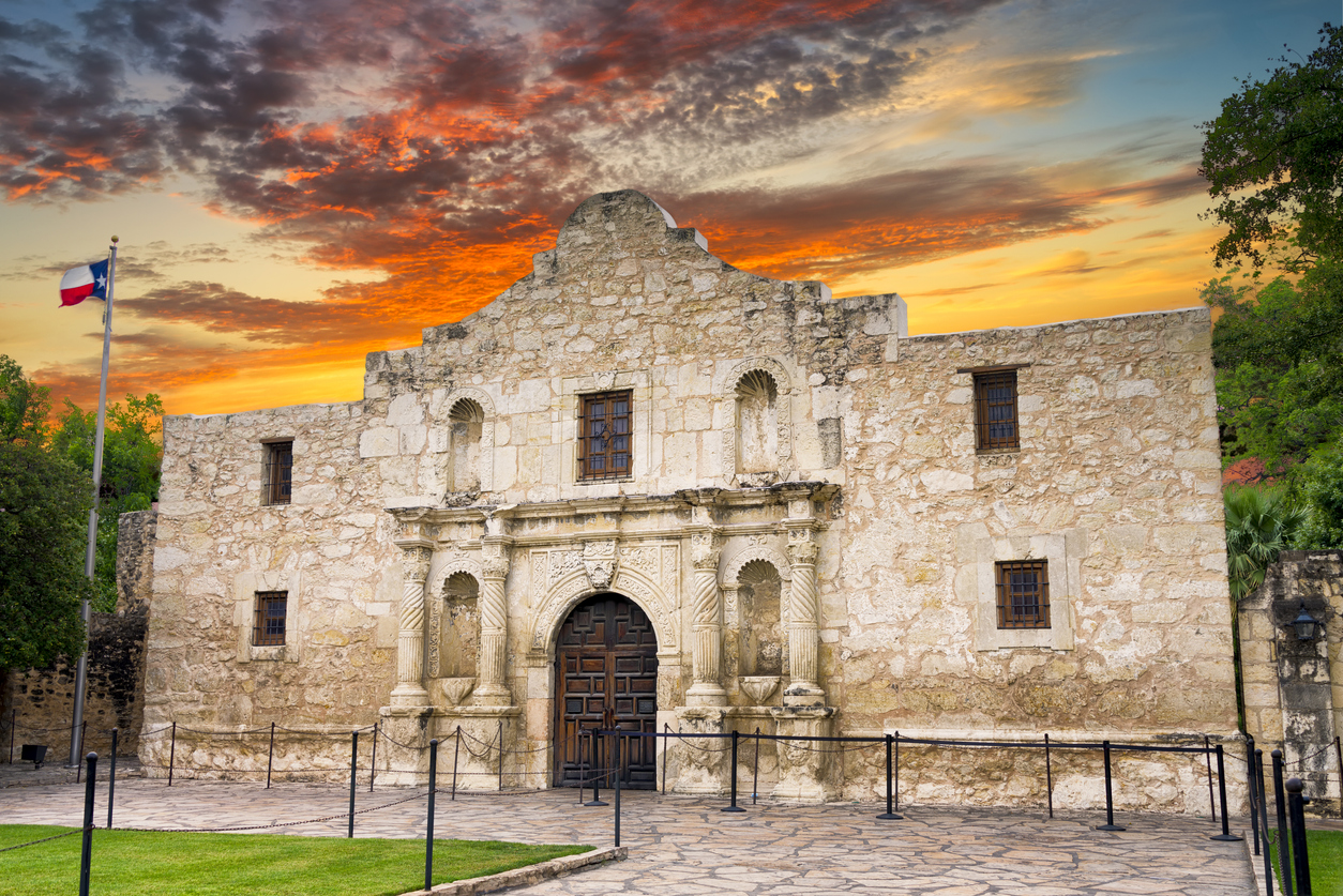 Die Fassade des Alamo in San Antonio, Texas, steht vor einem dramatischen Sonnenuntergangshimmel mit Texas-Flagge links.