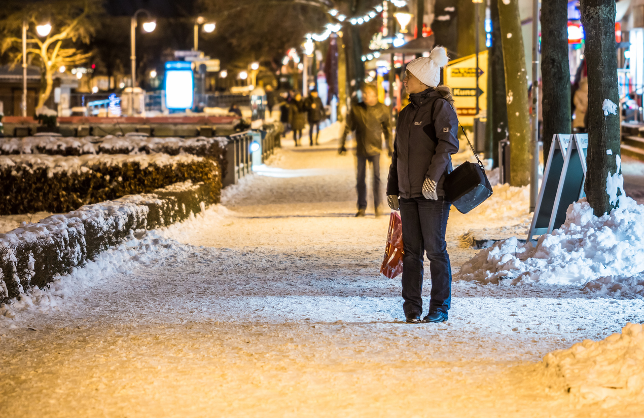 Frau mit Mütze und Einkaufstüte steht auf einem verschneiten, hell erleuchteten Gehweg in Warnemünde, während Passanten im Hintergrund vorbeigehen.