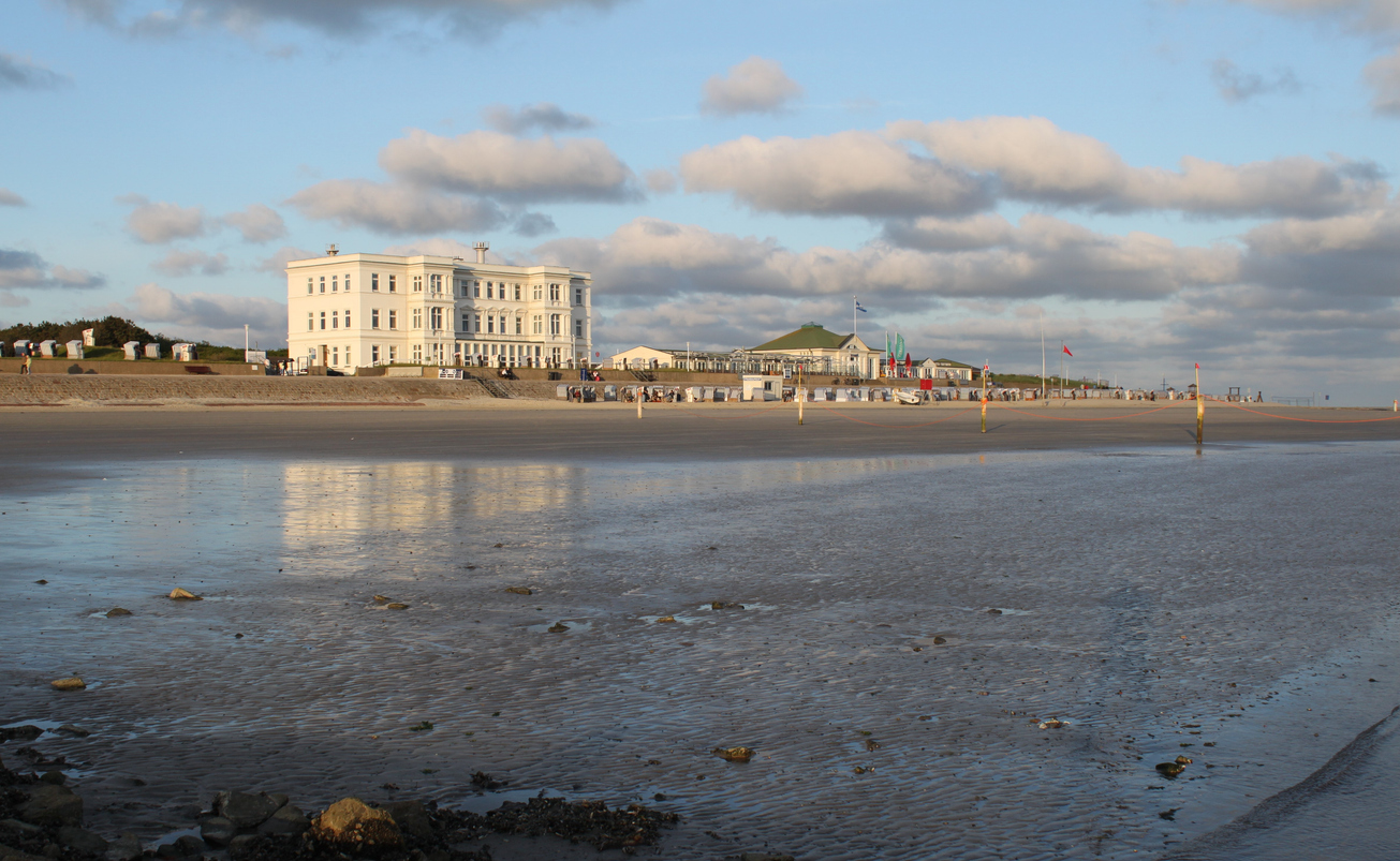 Blick über das Watt und den breiten Strand auf Norderney mit Promenade, Strandkörben und hellen Gebäuden im Abendlicht.