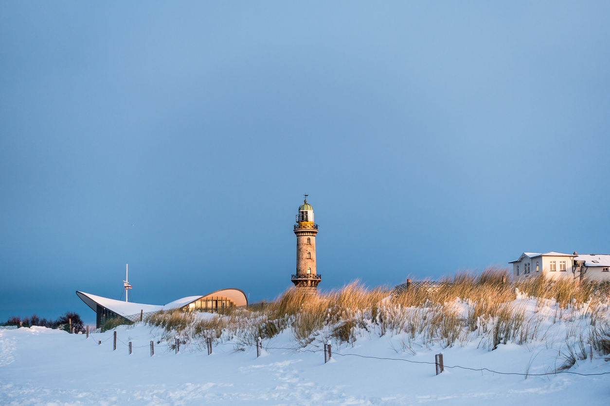 Verschneite Dünen mit Leuchtturm und Teepott in Warnemünde im warmen Licht der tiefstehenden Wintersonne.