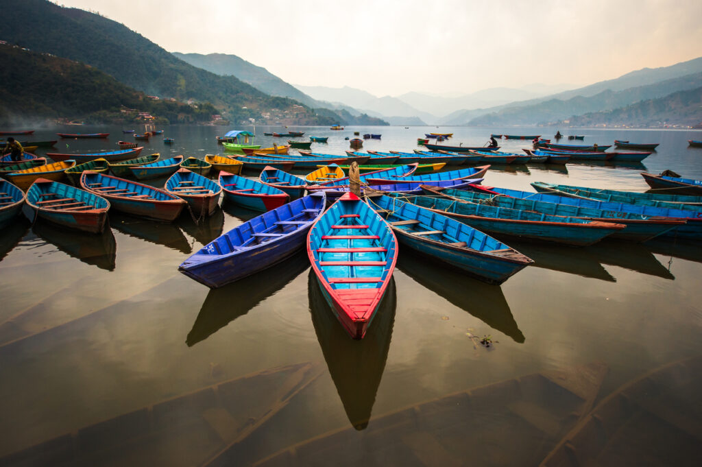 Viele farbenfrohe Holzboote liegen im ruhigen Wasser des Phewa-Sees bei Pokhara, umgeben von nebeligen Bergen.