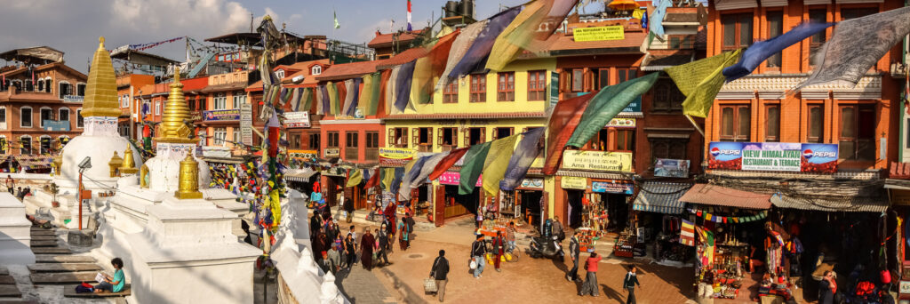 Straßenansicht in Kathmandu, Nepal, mit weißen Stupas, bunten Häusern, vielen Gebetsfahnen und Menschen.