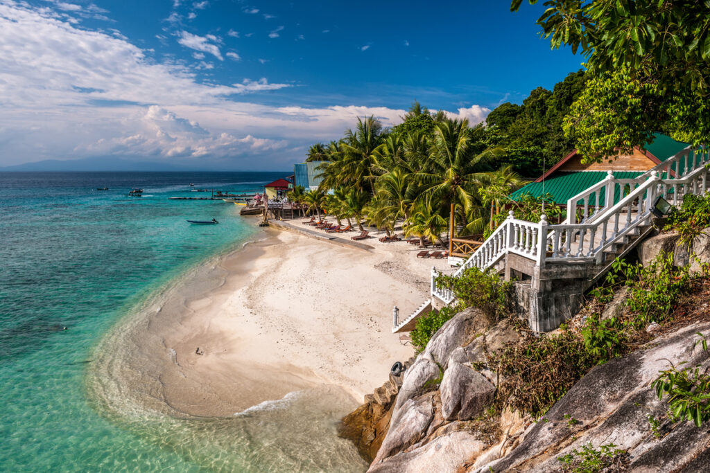An einem weißen Strand der Perhentian Islands stehen Palmen und Bungalows direkt über dem klaren Wasser.