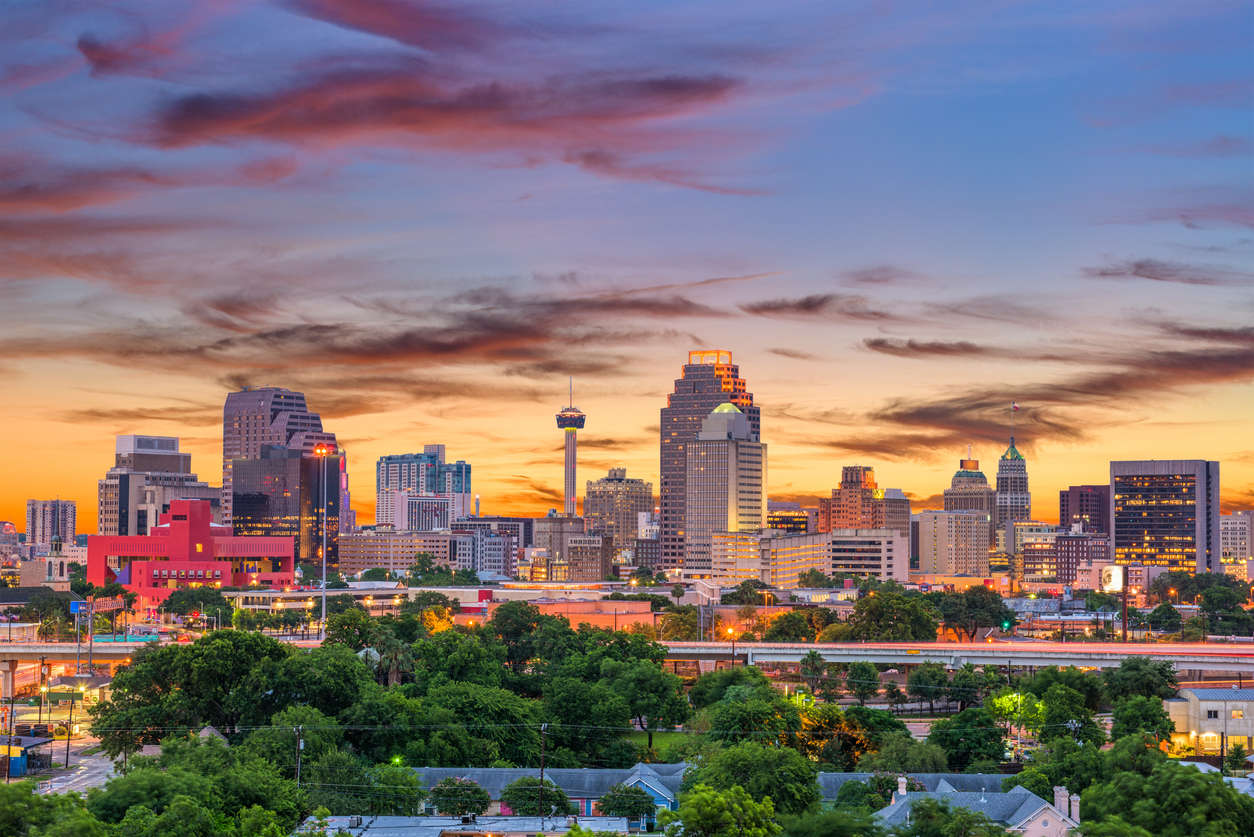 Panorama der Skyline von San Antonio, Texas, bei Sonnenuntergang mit beleuchteten Hochhäusern und buntem Himmel über einer grünen Vorstadt.
