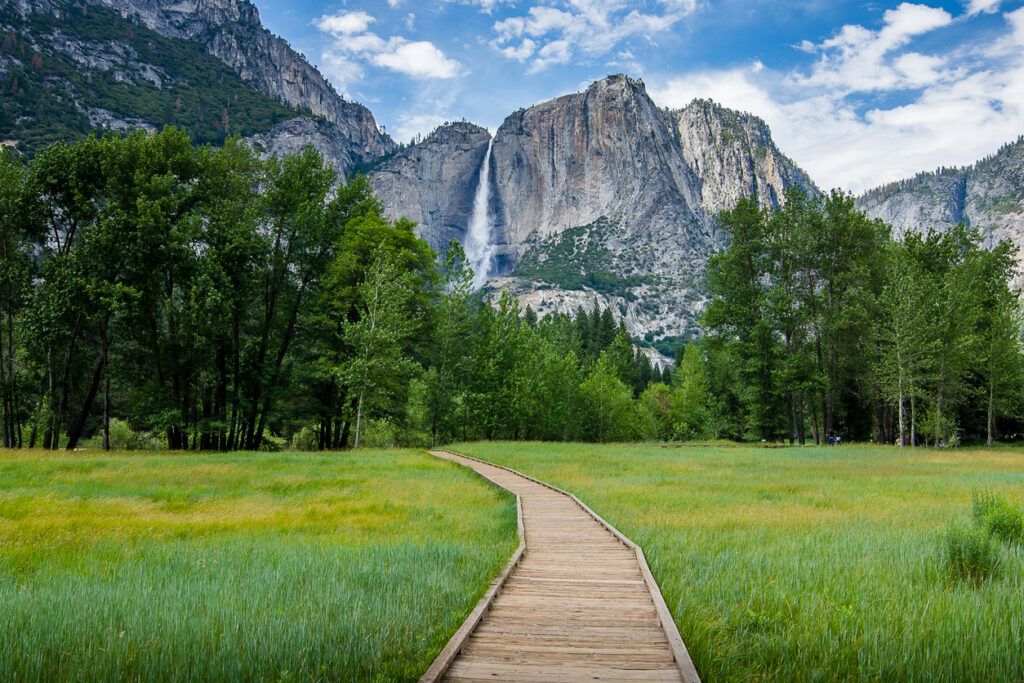 Ein Holzweg führt durch eine grüne Wiese auf Wasserfall und Granitfelsen im Yosemite Nationalpark zu.