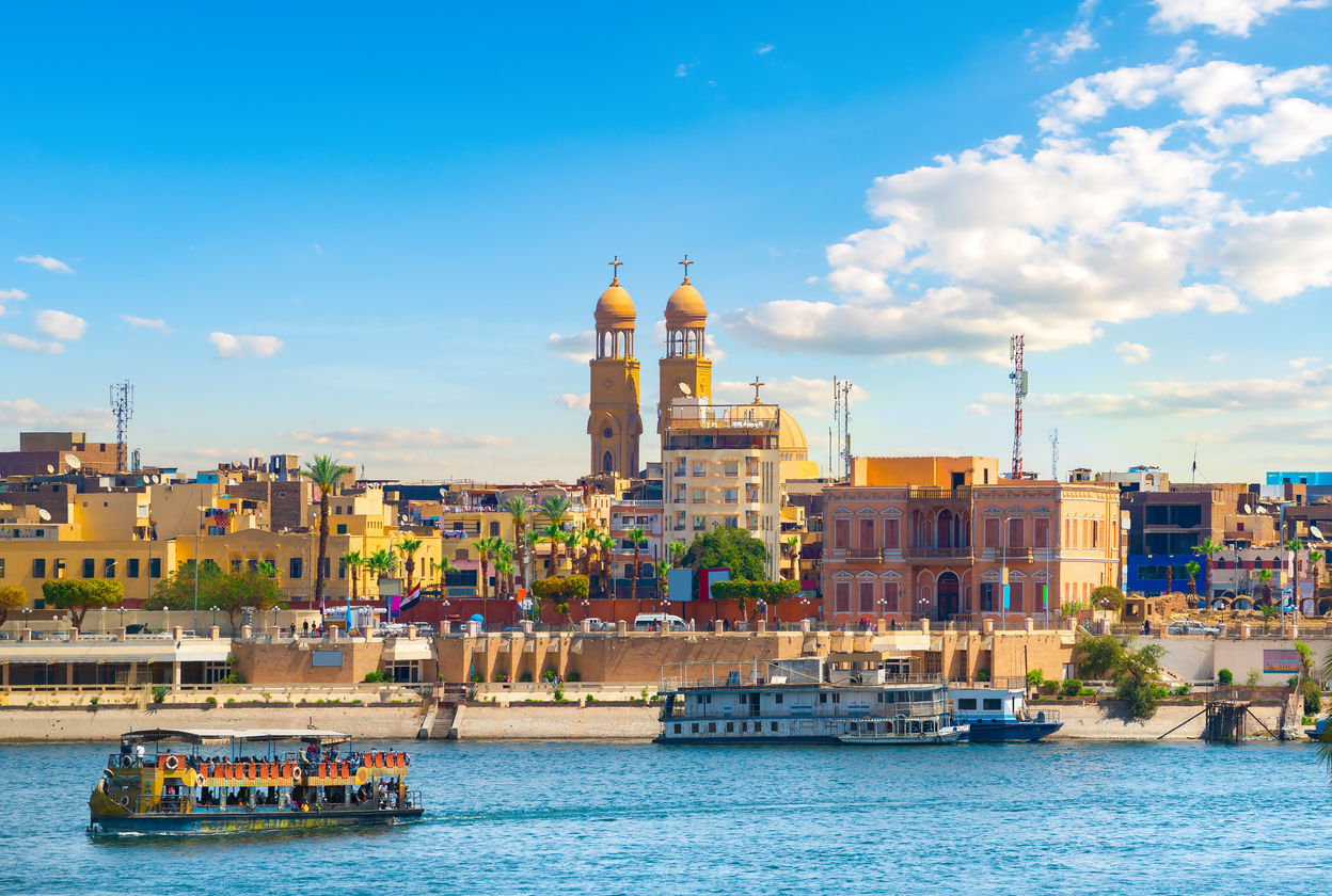 Am Nil fahren Boote vorbei, dahinter stehen bunte Gebäude und markante Türme unter blauem Himmel.