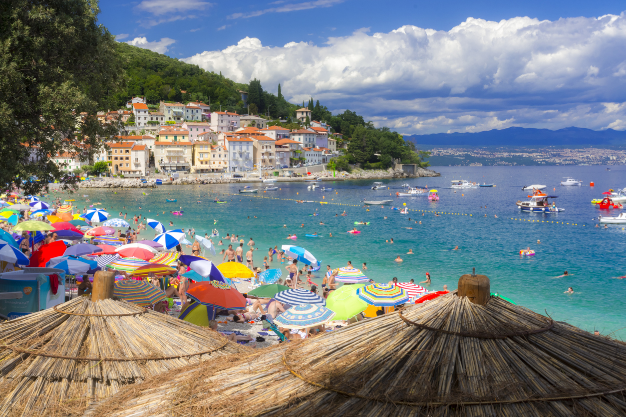 Volle Strandbucht in Kroatien mit bunten Sonnenschirmen, Badegästen, Booten und Häusern am Hang.
