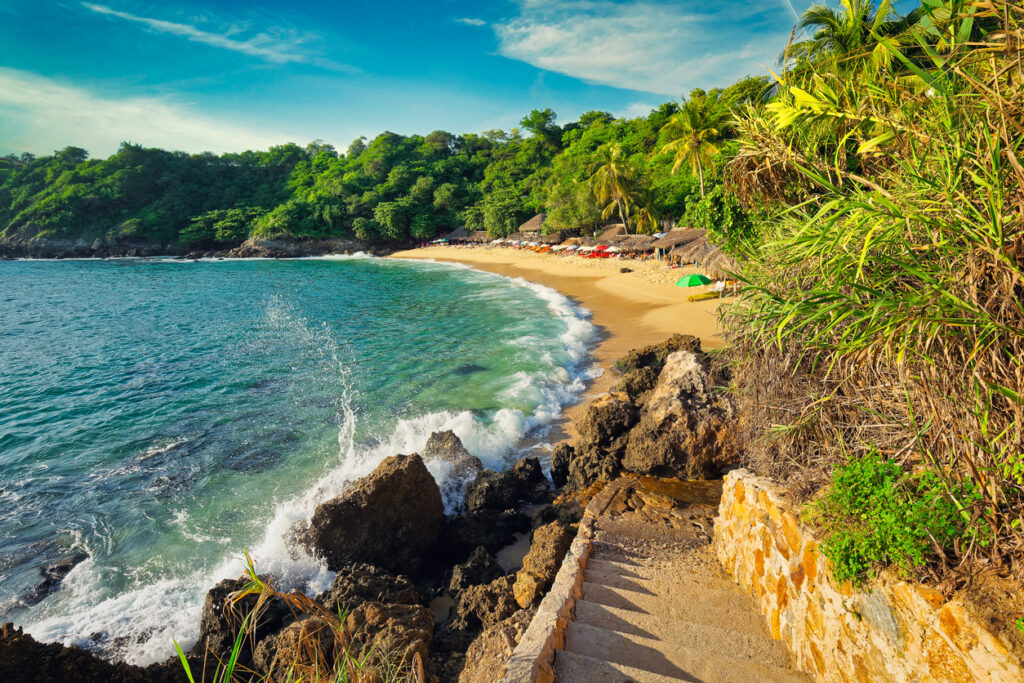 Blick über eine tropische Bucht in Puerto Escondido mit goldenem Strand, Palmen, Felsen und türkisblauem Meer.